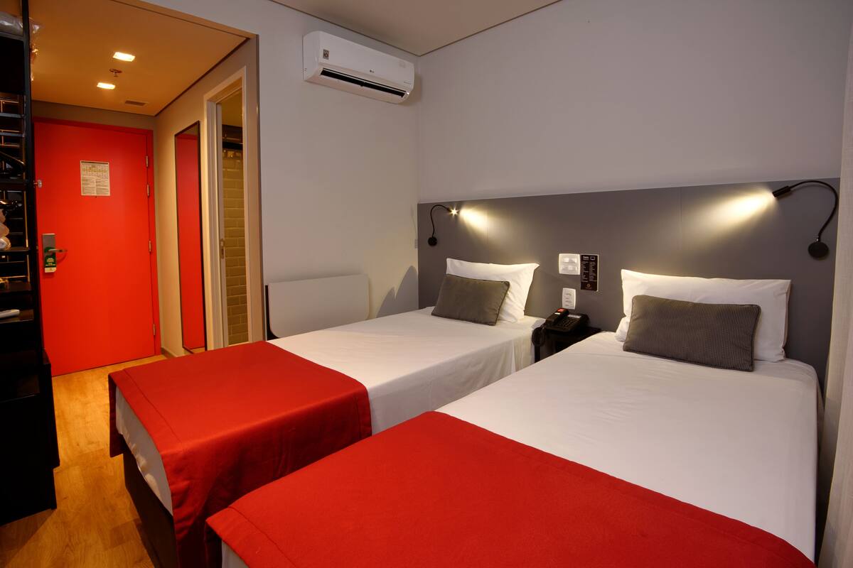 Ein Hotelzimmer mit zwei Einzelbetten, beide mit roter Bettdecke und weißen Laken. Es gibt eine rote Tür, eine weiße Klimaanlage an der Wand sowie einen Nachttisch mit einer Lampe zwischen den Betten.