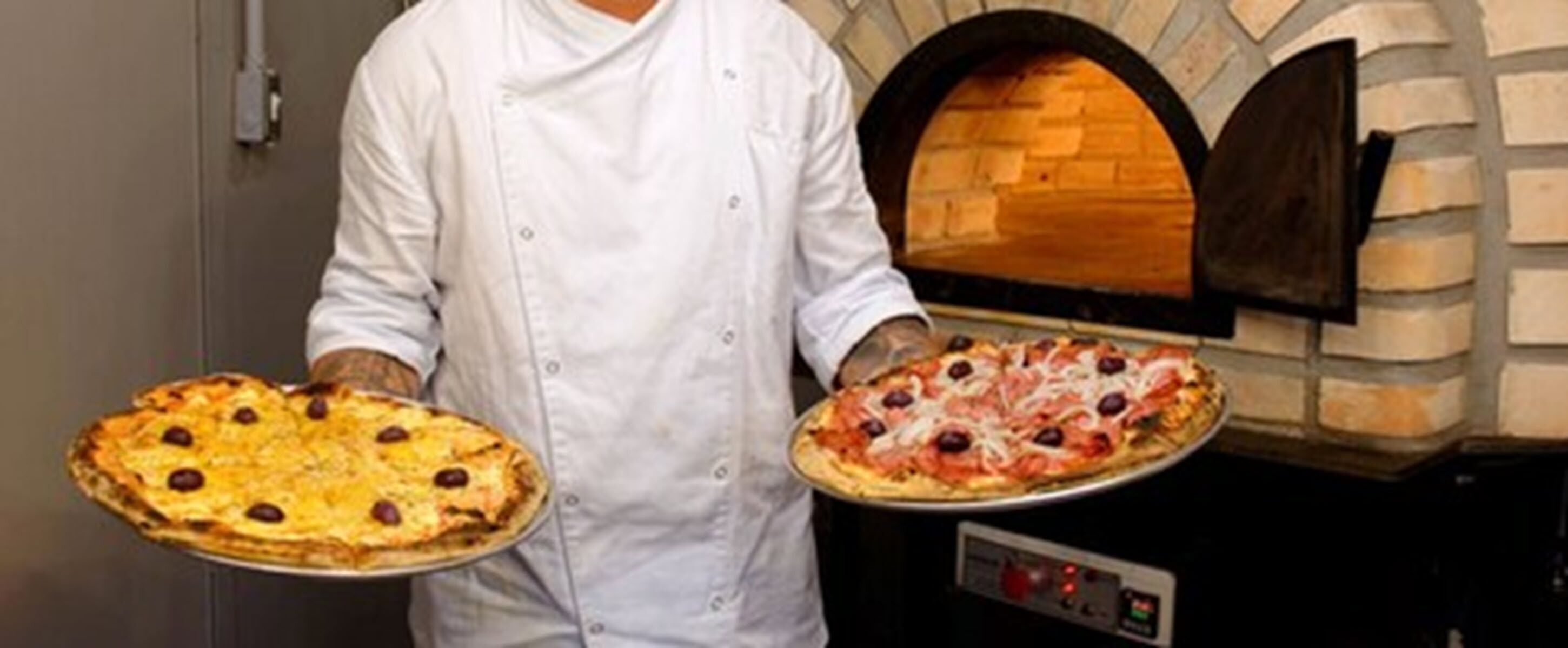 Un chef sostiene dos pizzas, una con aceitunas y otra con queso, frente a un horno de ladrillo.