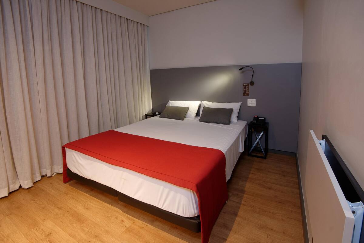 Ein Hotelzimmer mit Bett, Nachttisch, Lampe und einem Fenster mit Vorhängen.