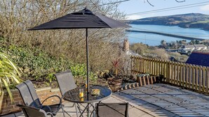 An outdoor patio with a table, chairs, and an umbrella, overlooking a body of water and hills. - Picked for its wonderful character and charm and stunning sea views to enjoy. (Goodwick)