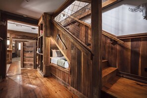 Una escalera de madera con una cama empotrada y estanterías, que conduce a una habitación con una ventana y una puerta al fondo.