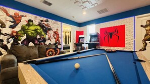 Una mesa de billar con una bola, un sofá con cojines y una pared con murales de superhéroes.