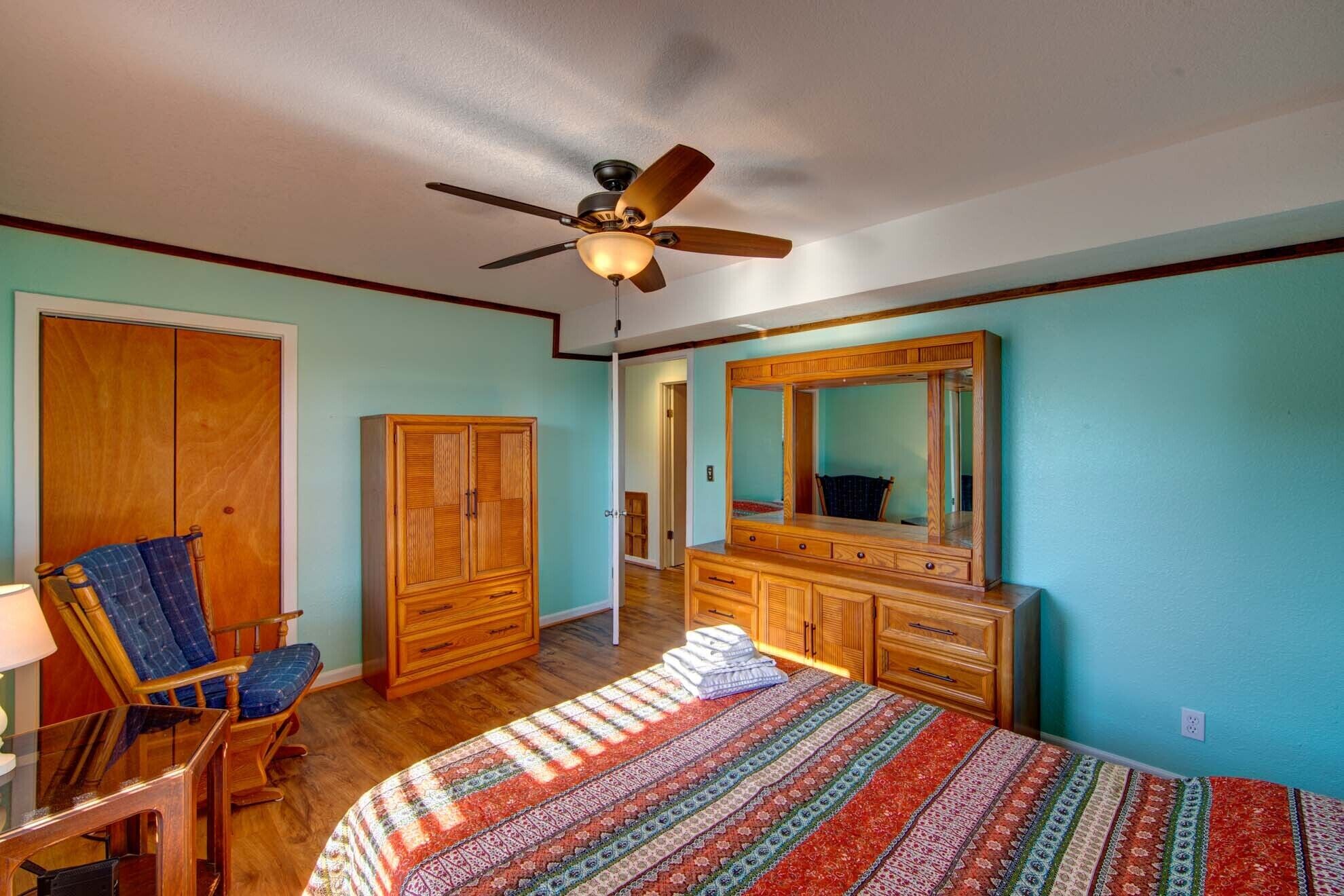 Ein Schlafzimmer mit einem Bett, einem Holzschrank, einem Schminktisch, einem Sessel und einem Deckenventilator.