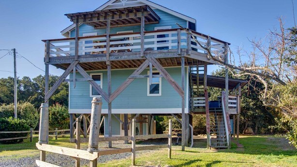 A two-story wooden structure with a balcony, surrounded by trees and a gravel path. - Surf Chalet w/ hot tub and kayaks (Avon)