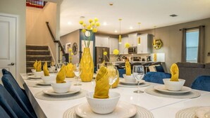 Ein Esszimmer mit einem langen Tisch, gedeckt für eine Mahlzeit, eine Küche im Hintergrund und eine Treppe, die zu einer anderen Ebene führt.