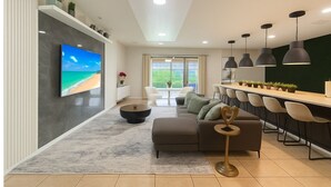 A modern living room with a flat-screen TV, a sofa, a coffee table, and a dining area with chairs and pendant lights.