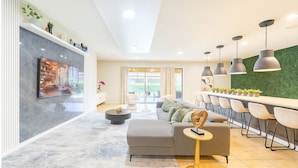 A modern living room with a sofa, coffee table, and a TV. There is a dining area with chairs and pendant lights.