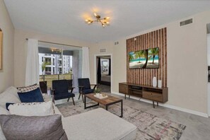 A living room with a sofa, chairs, a coffee table, a TV on a wooden wall, and a view through a glass door.