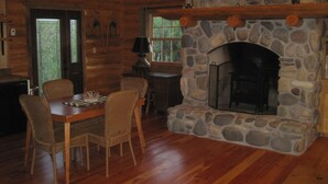 A rustic cabin interior with a stone fireplace, wooden dining table, wicker chairs, and a stone wall.