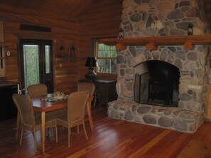 A rustic cabin interior with a stone fireplace, wooden dining table, wicker chairs, and a stone wall.