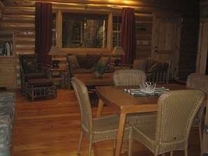 A cozy log cabin interior with a wooden dining table, wicker chairs, a sofa, and a window with curtains.