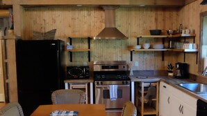A rustic wooden interior with a dining table, wicker chairs, a black refrigerator, a stove, and a sink.