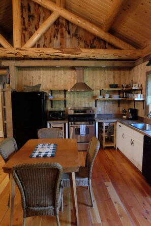 A rustic wooden interior with a dining table, wicker chairs, a black refrigerator, a stove, and a sink.