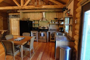 A log cabin kitchen with a dining table, chairs, a stove, oven, sink, and a trash can.