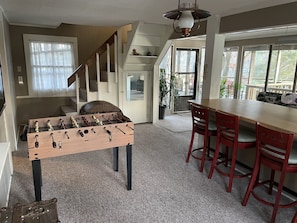 A cozy room with a wooden foosball table, red bar stools, a staircase, and large windows offering a view of the outdoors.