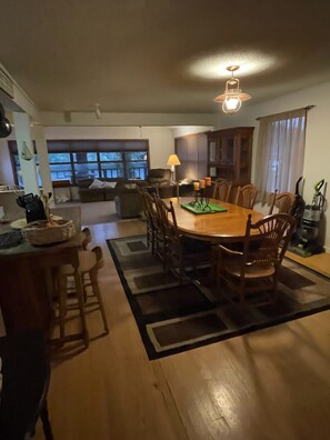 A spacious dining area with a wooden table and chairs, a lamp, and a view of the outdoors through large windows.