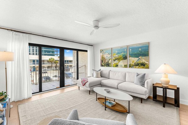 A modern living room with a sectional sofa, a coffee table, a ceiling fan, a large window with a view of a building and a balcony, and a painting on the wall. - Beachfront Condo with Stunning Ocean View with Heated Pool (Cocoa Beach)