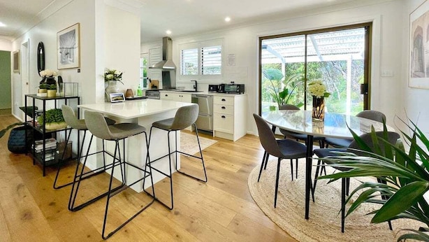 Een moderne keuken met witte kasten, een centraal eiland, barstoelen en een eettafel met stoelen.