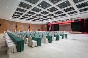 A conference room with rows of tables and chairs, a stage with a red curtain, and a high ceiling with a grid pattern. - Garden Hotel (Yilong)