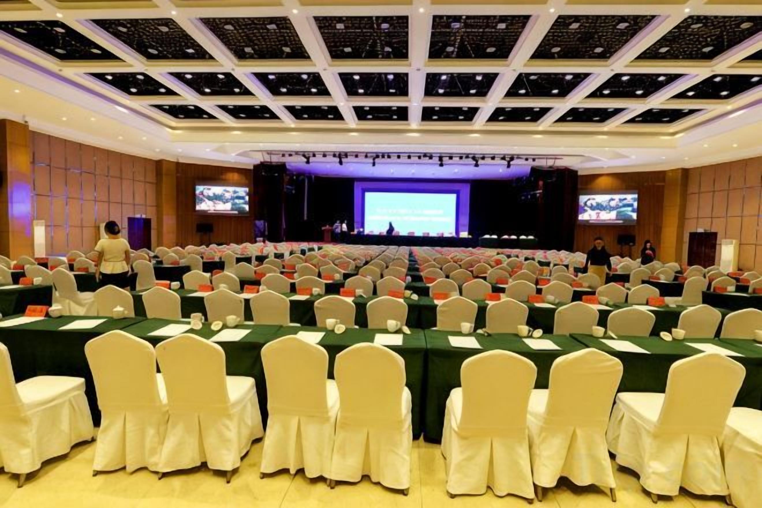 A conference room with rows of tables and chairs, a stage with a large screen, and a television.