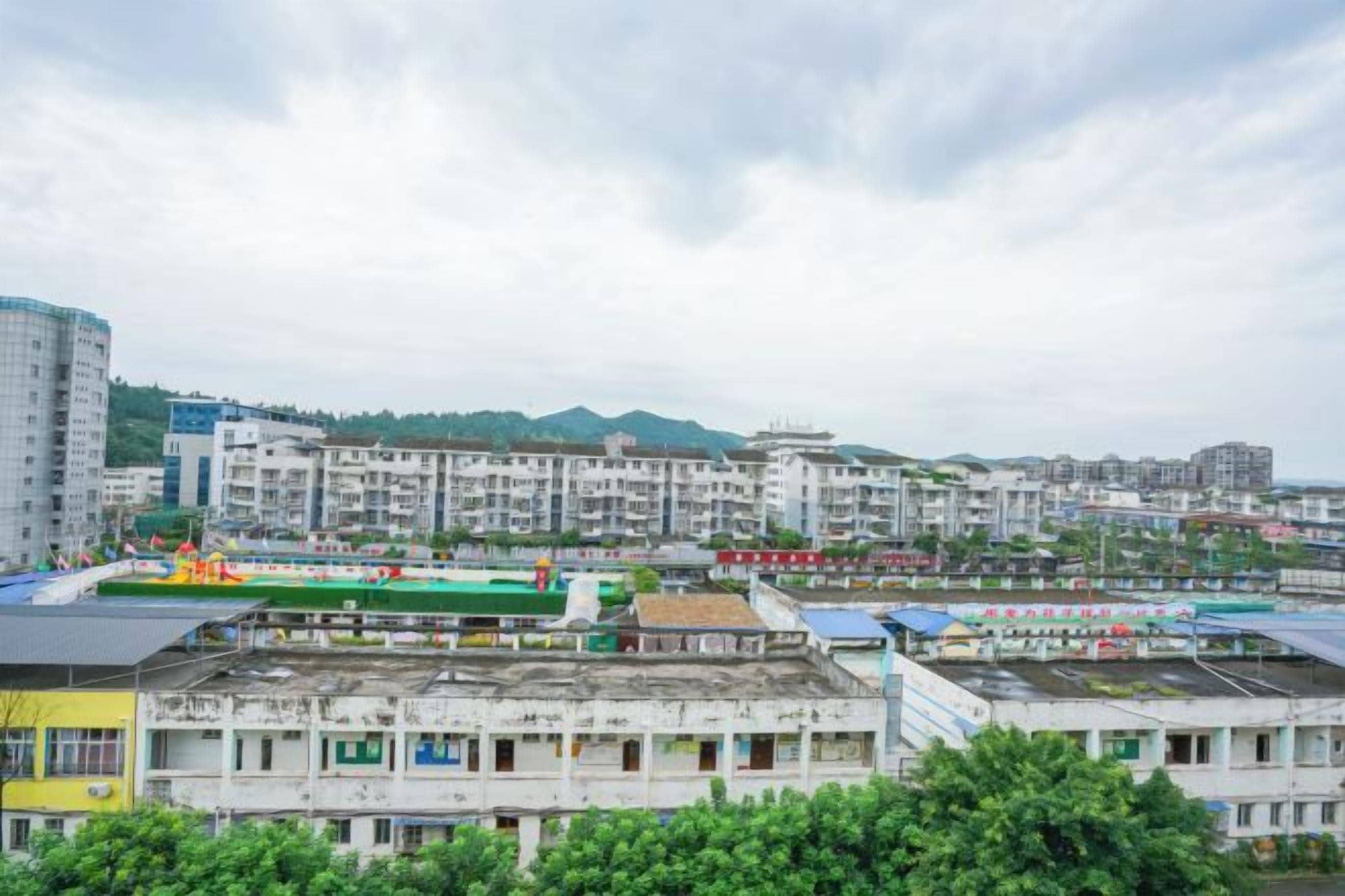 Um edifício de vários andares com telhado plano, rodeado por vegetação e outros edifícios.