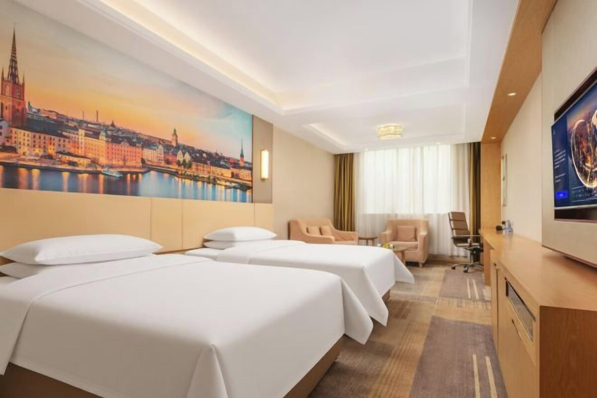 Ein Hotelzimmer mit zwei Betten, einem Schreibtisch, einem Sessel, einem Fernseher und einem Stadtbild.
