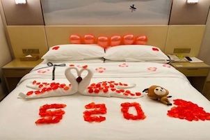Habitación de hotel con una cama decorada con pétalos de rosa y un arreglo con forma de cisne, un peluche, y el número "520" escrito con pétalos.