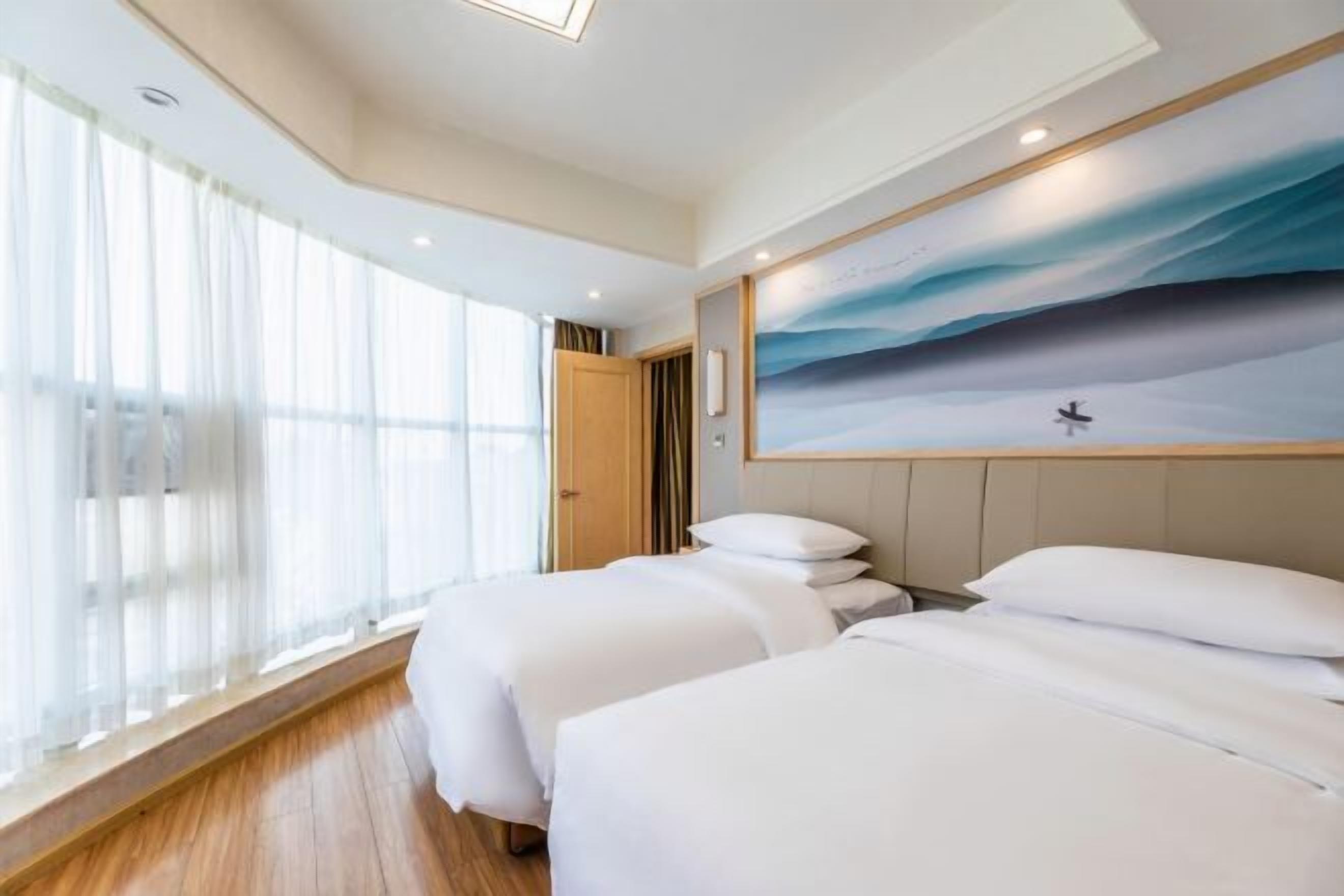 A hotel room with two beds, a large window with sheer curtains, a painting of a seascape on the wall, and a wooden floor.