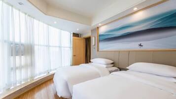A hotel room with two beds, a large window with sheer curtains, a painting of a seascape on the wall, and a wooden floor.