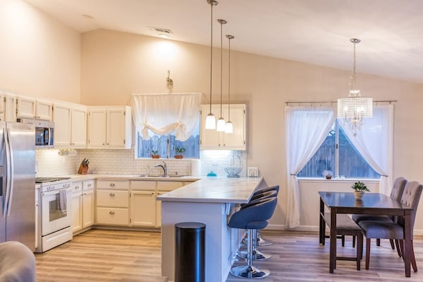 A modern kitchen with white cabinets, a central island, pendant lighting, and a dining area with a table and chairs.