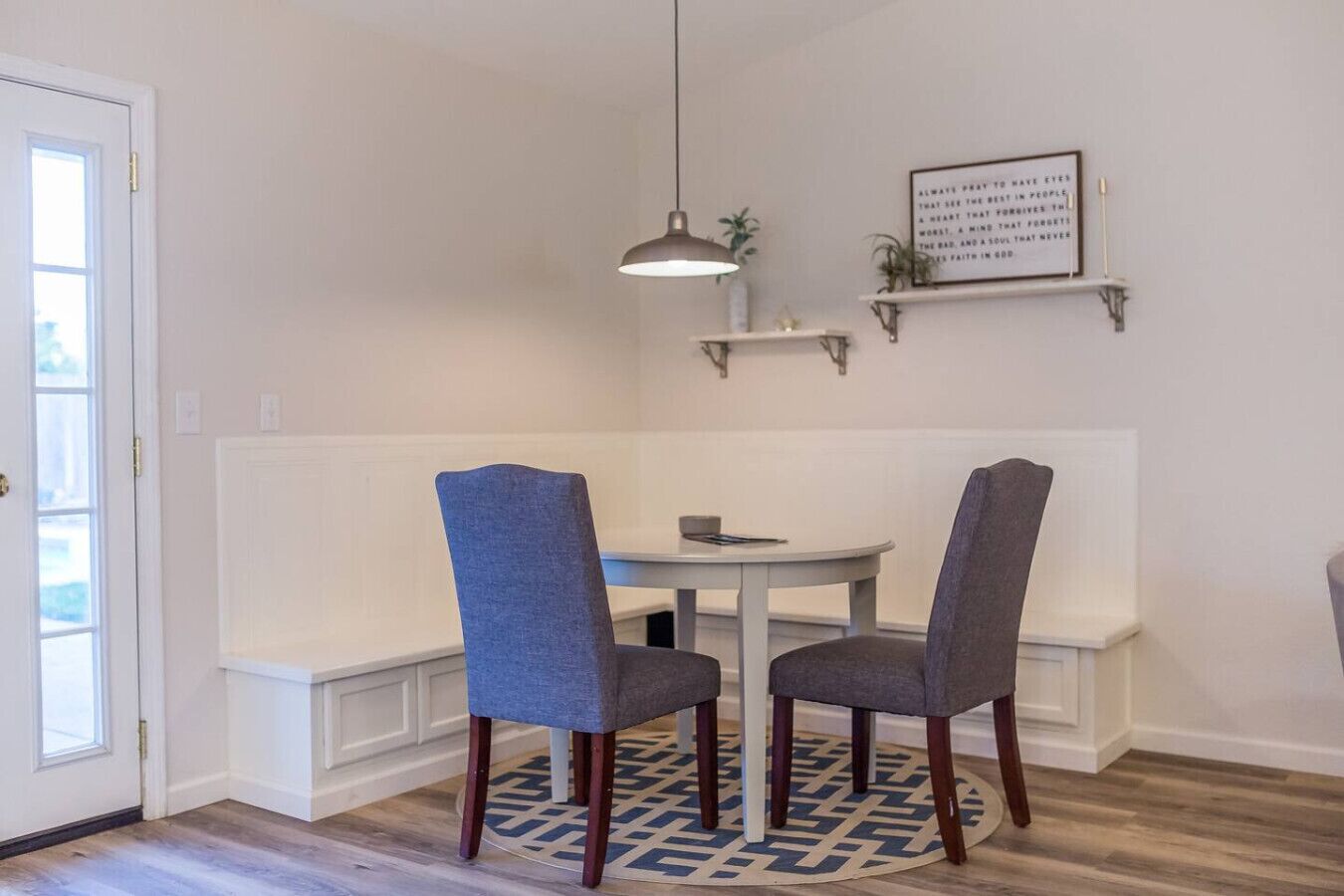 Un coin repas comprenant deux chaises, une table, une suspension, une étagère fixée au mur avec une plante, et une porte.