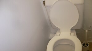 Des toilettes blanches avec un abattant fermé, dans une salle de bain aux murs blancs et au sol carrelé.