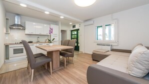 Et moderne køkken med hvide skabe, et spisebord med stole, en sofa og et vindue med udsigt til grønne omgivelser.