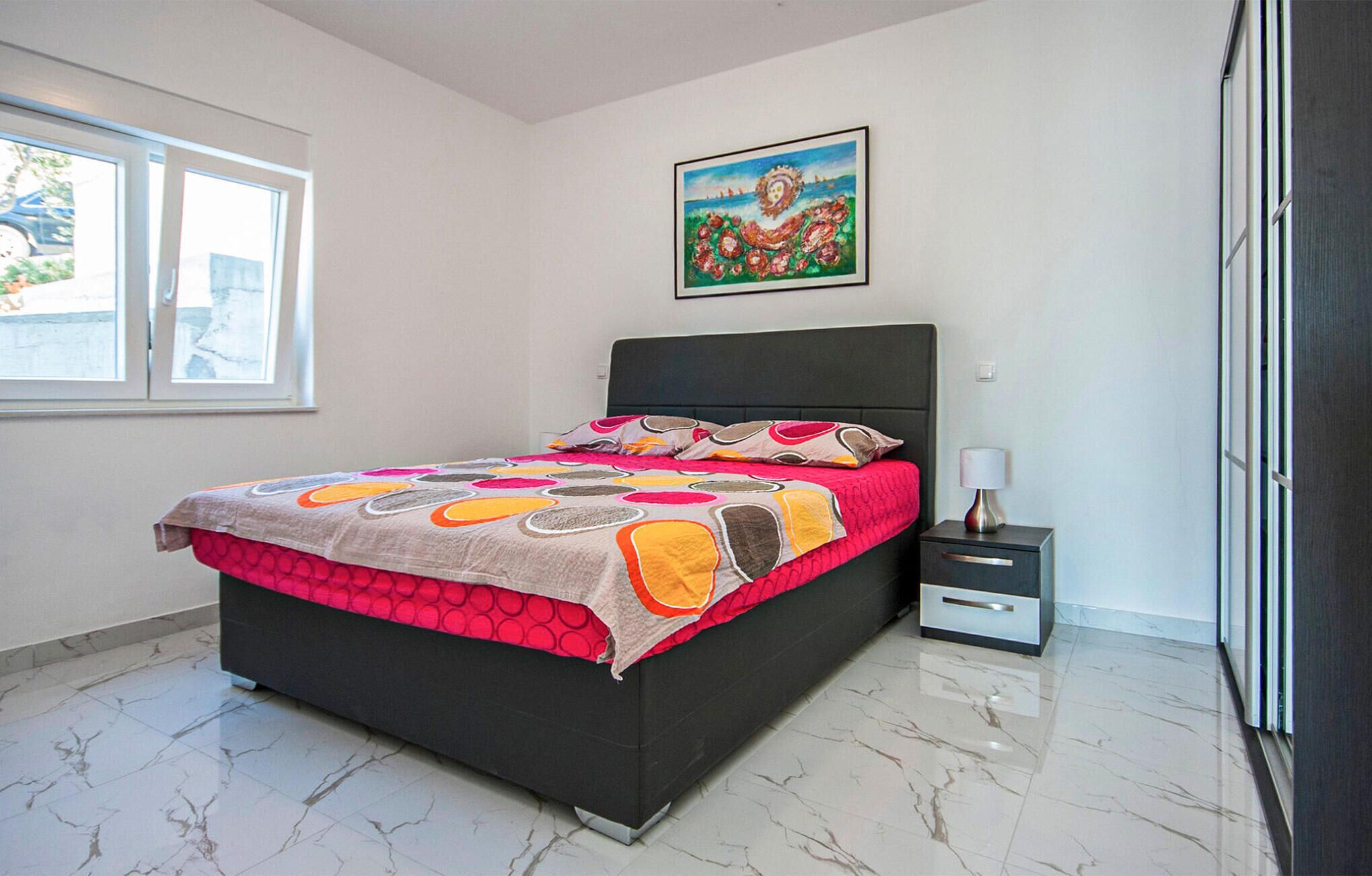 A bedroom with a bed, a nightstand, a lamp, a painting, and a window.