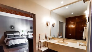 A hotel room with a bed, bedside table, lamp, and a large mirror above the sink.