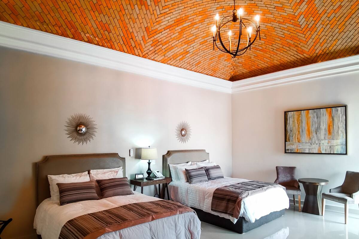 Une chambre d’hôtel avec deux lits, un lustre, un bureau avec une lampe et un tableau au mur.
