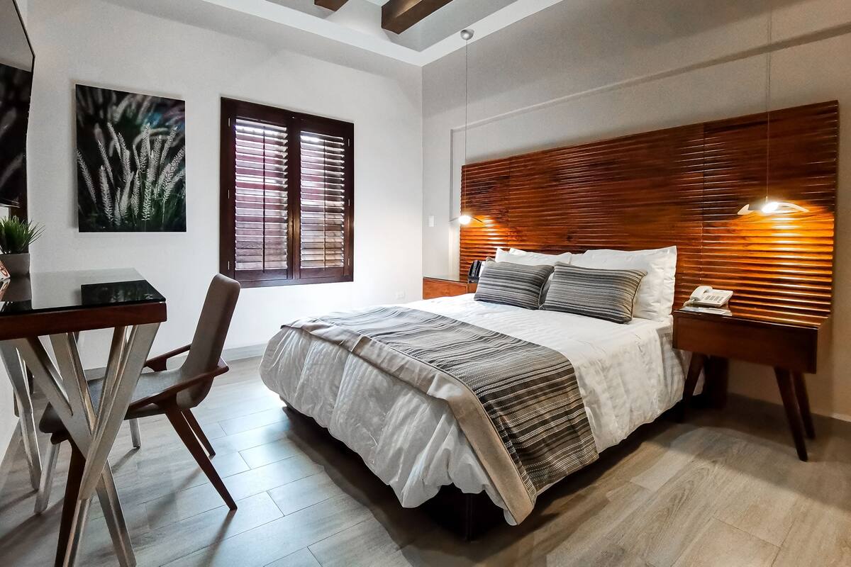 Une chambre à coucher comprenant un lit, une table de chevet, une chaise, un bureau et une œuvre d’art accrochée au mur.