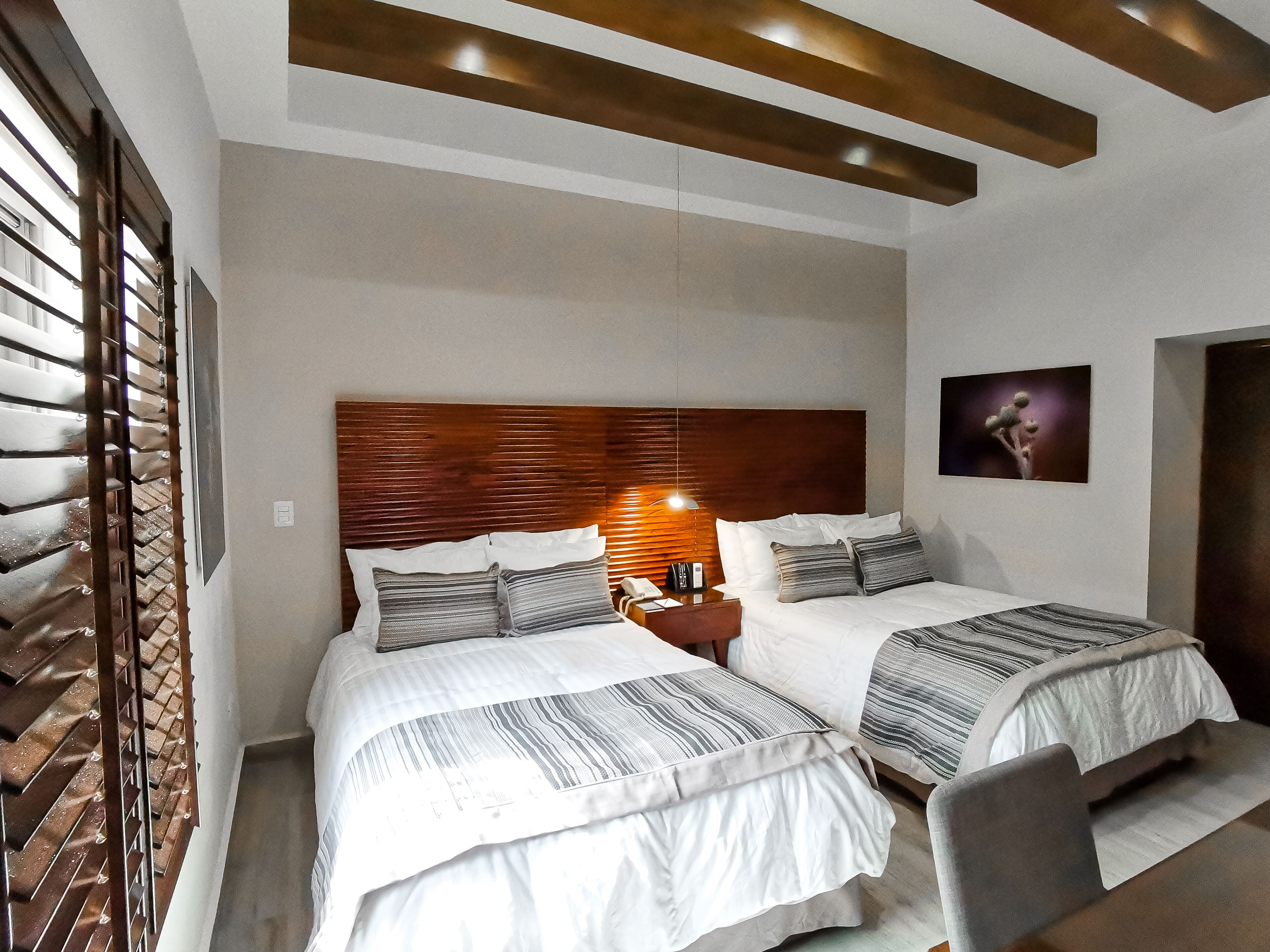 Habitación de hotel con dos camas, cabecero de madera, mesita de noche con lámpara y un cuadro en la pared.