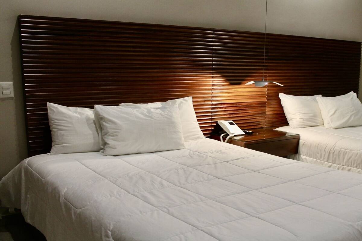 Une chambre d’hôtel avec deux lits, une tête de lit en bois, du linge de lit blanc, des tables de chevet et un téléphone.
