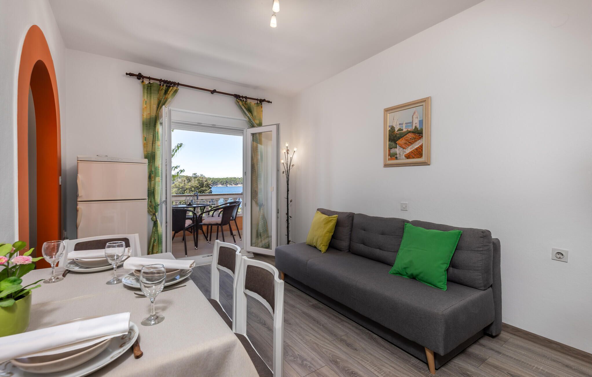 Un coin repas avec une table dressée pour deux personnes, un canapé gris avec des coussins verts et un tableau accroché au mur.