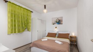 Une chambre à coucher comprenant un lit, une table de chevet, une lampe fixée au mur, une porte et une fenêtre avec des rideaux légers.