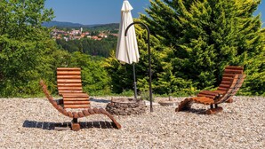 Two wooden lounge chairs with a stone fire pit and a white umbrella in a garden setting with a view of a village and mountains.
