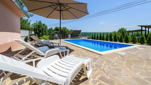 A swimming pool with white lounge chairs and an umbrella, surrounded by stone flooring and greenery. - Awesome apartment in Podpican (Podpican)