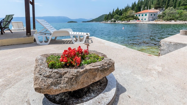 Un vaso in pietra con fiori rossi su una terrazza in cemento, vicino all'acqua, con poltrone relax e un edificio sullo sfondo.