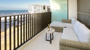 A balcony with a white sofa, a small round table with a bottle and glass, and a view of the beach and buildings. - Nice apartment in Oostende (Oostende)
