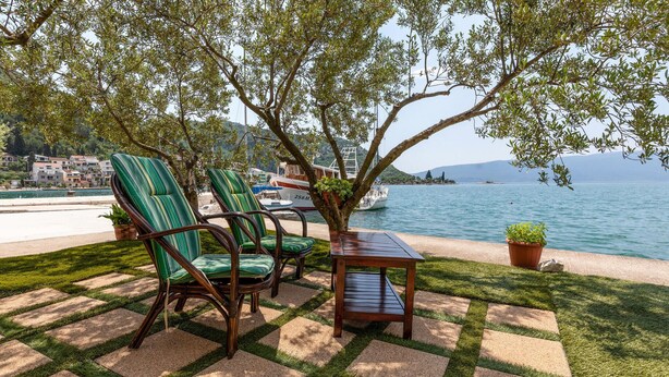 Une terrasse avec deux chaises à rayures vertes et une table en bois, donnant sur une étendue d’eau où l’on aperçoit des bateaux et des bâtiments au loin.