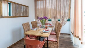 Un coin repas avec une table en bois dressée pour quatre personnes, entourée de chaises en osier avec des coussins rouges. Un vase rempli de fleurs violettes est posé sur la table.