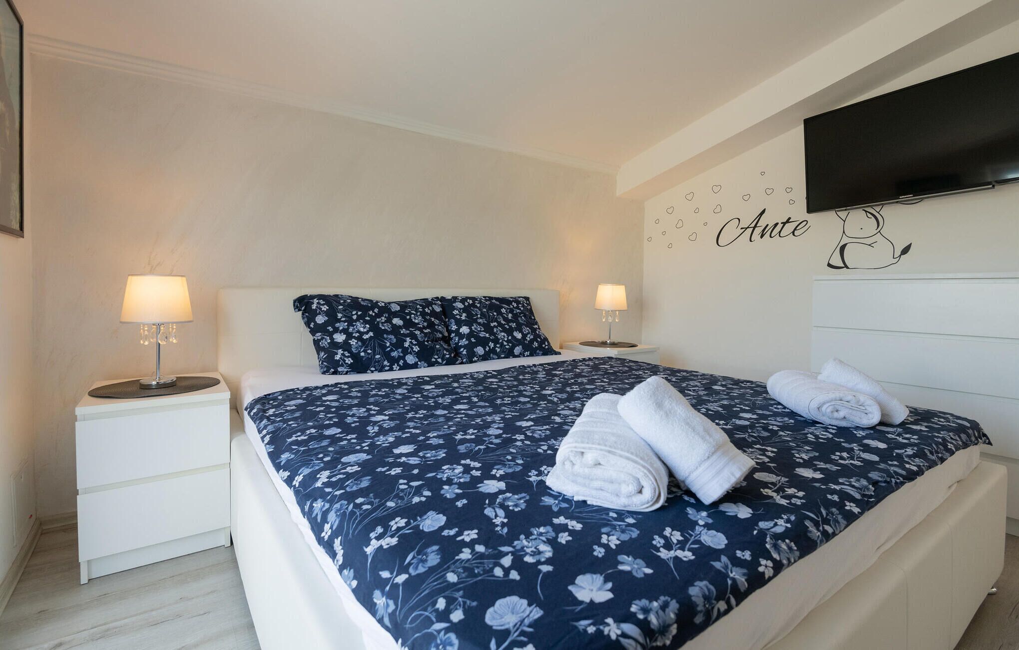 A bedroom with a bed, two bedside tables, a TV mounted on the wall, and a wall decoration with the word "Ante".