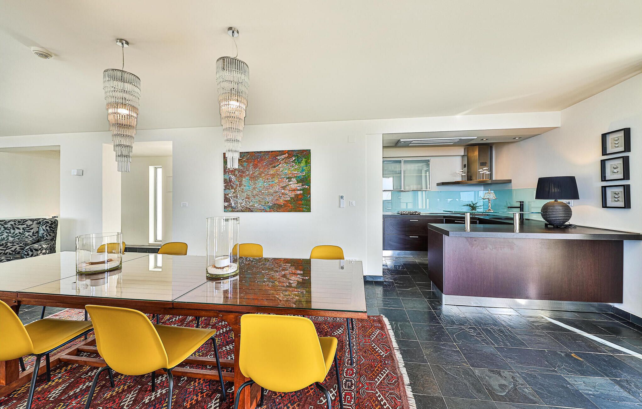 A modern dining area with a glass table, yellow chairs, and a chandelier.