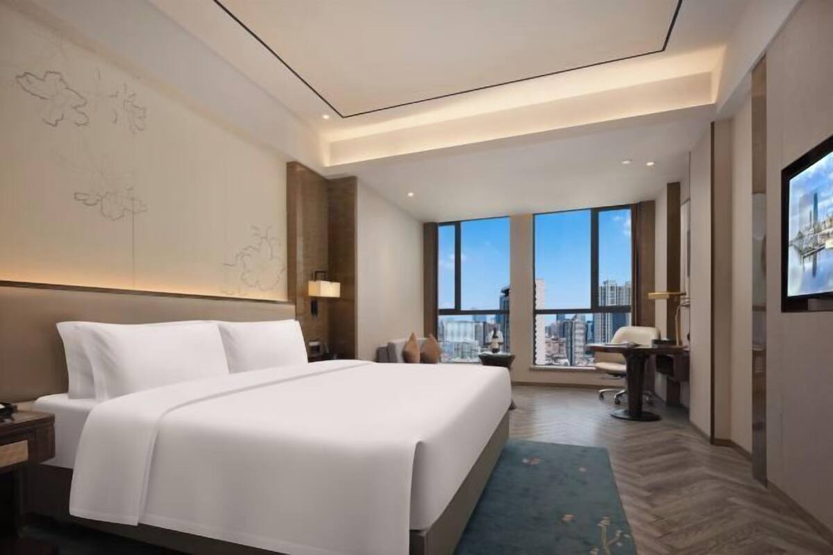 Ett modernt hotellrum med en stor säng, ett skrivbord och utsikt över staden genom stora fönster.